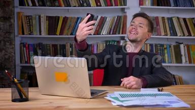 有魅力的男学生在图书馆里坐在笔记本电脑前打电话自拍